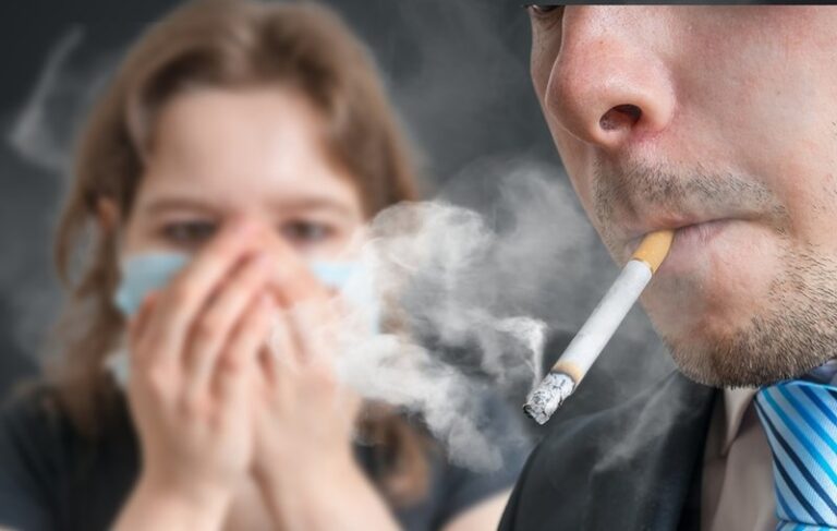 a person is smoking cigarette and a girl in background wearing mask and both hand on her nose to save herself from the cigarette smoke.