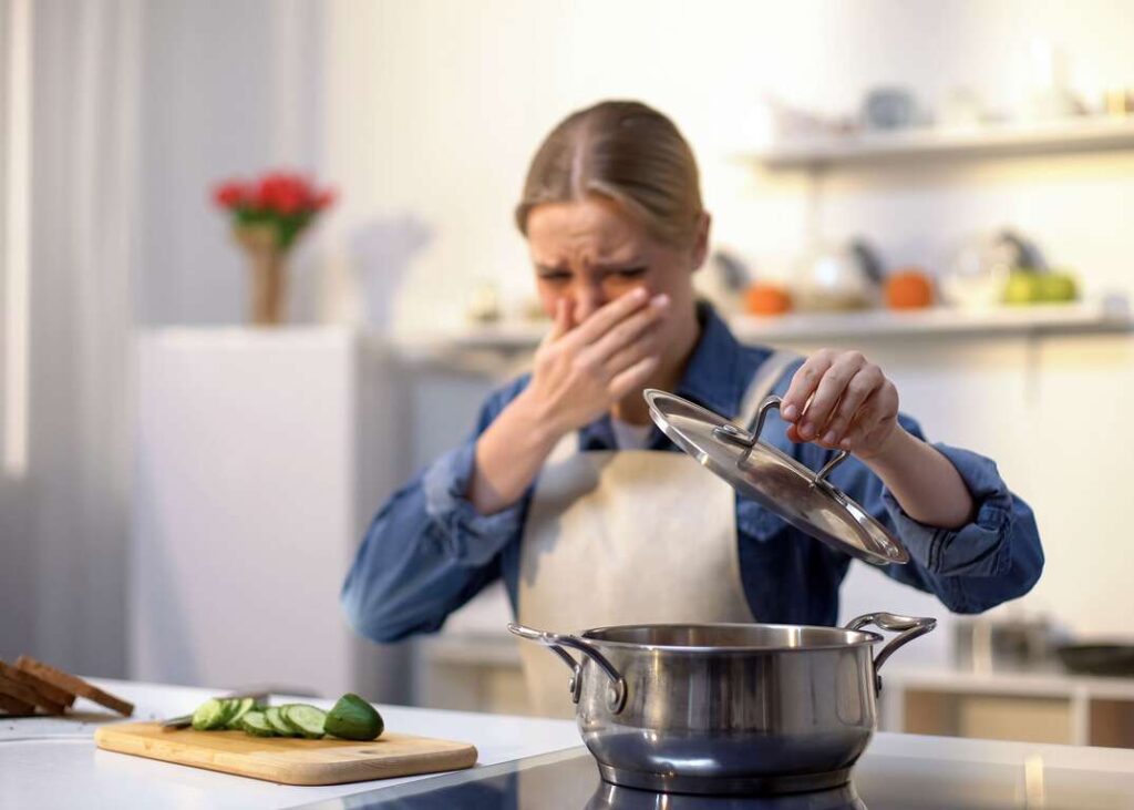 A girl lifts the lid of the pot and puts her hand on her nose as if she is smelling something bad.